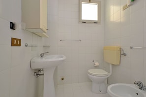 A bathroom with a sink, toilet, and bidet, white tiled walls, and a window. - Adorable Apartment With Pool (Lignano Sabbiadoro)