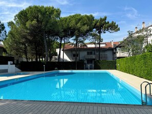 A swimming pool with tiled edges, surrounded by trees and a building with a red-tiled roof. - Welcoming Apartment for Groups (Lignano Sabbiadoro)