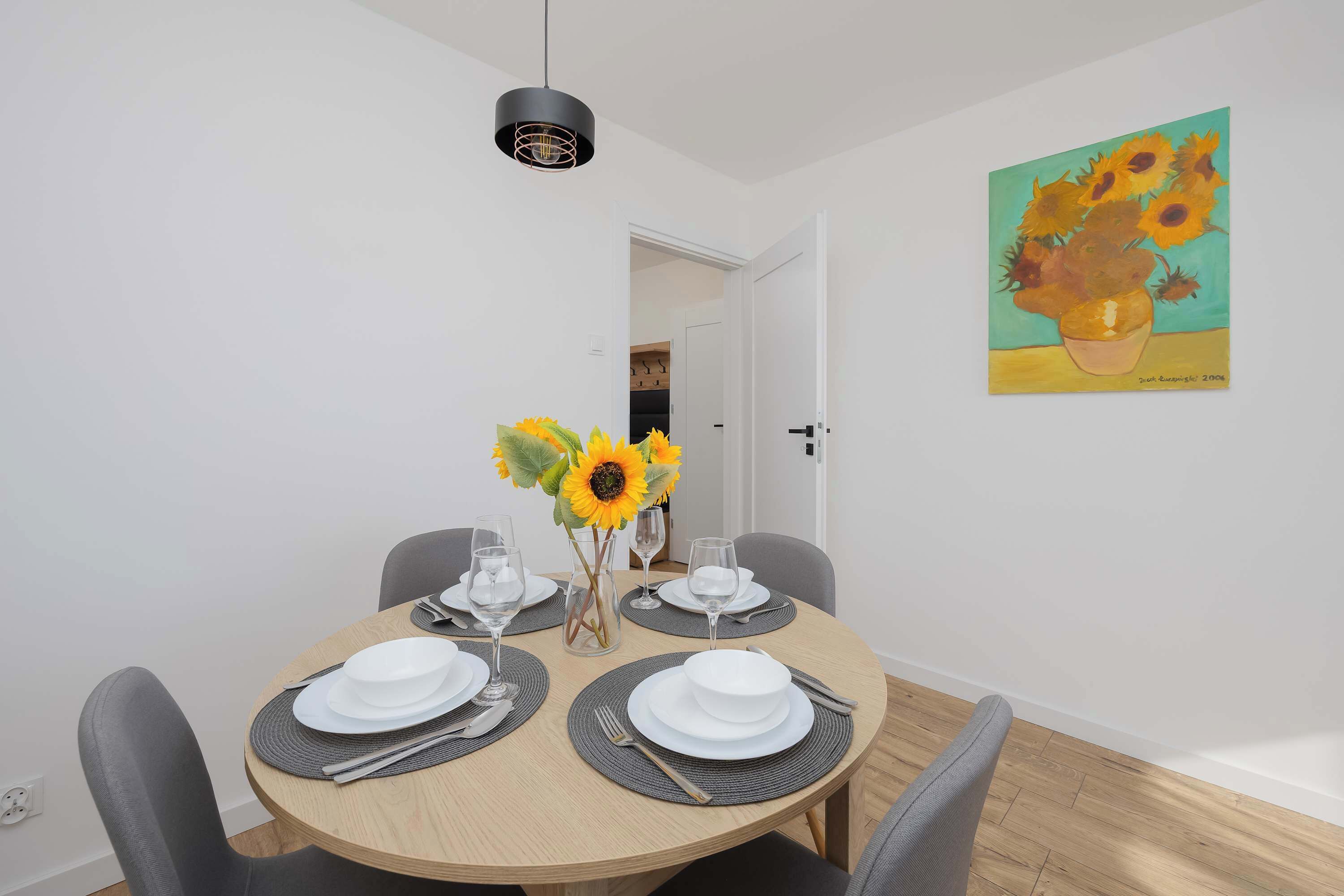Un comedor con una mesa redonda dispuesta para cuatro personas, un jarrón con girasoles y un cuadro en la pared.
