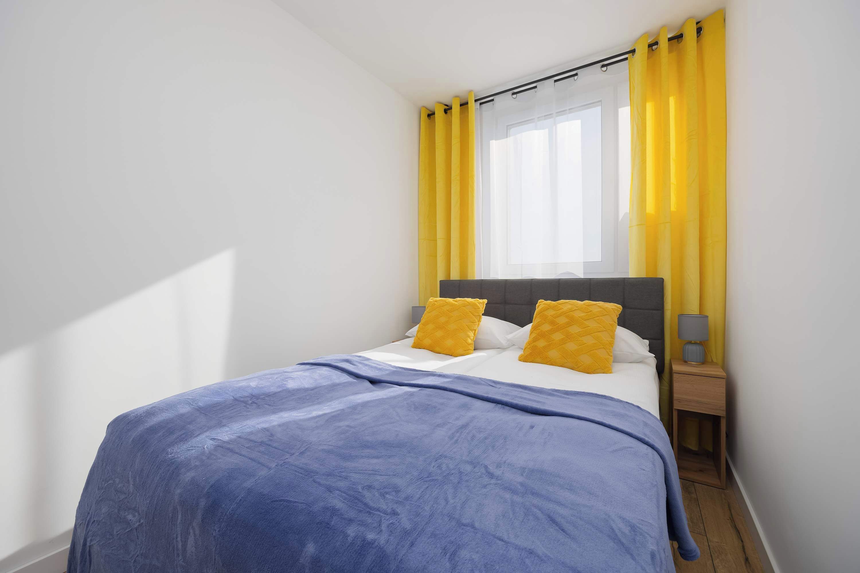 A bedroom with a bed, a nightstand, a window with yellow curtains, and a headboard.