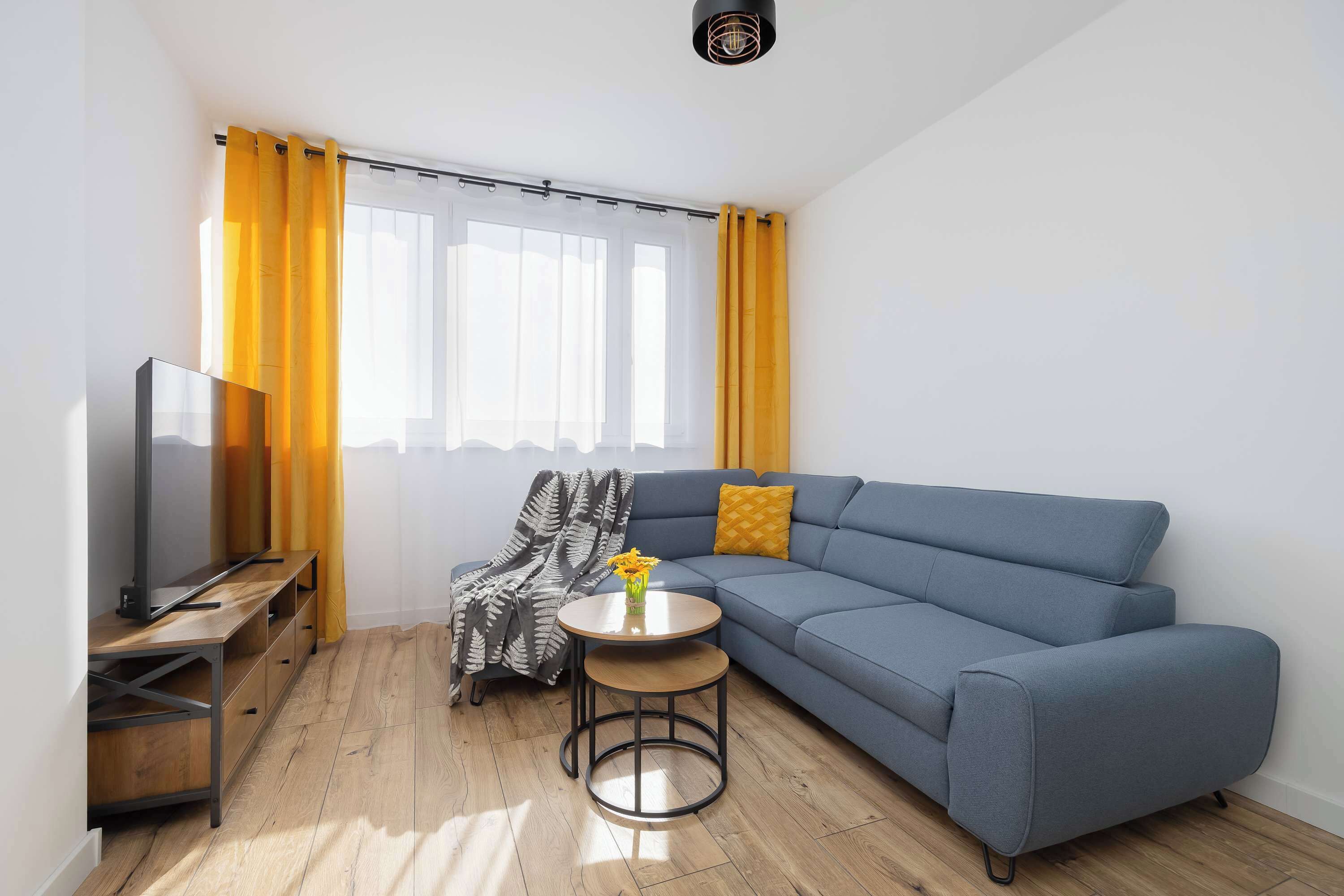 A modern living room with a blue sofa, a TV on a wooden stand, a small round table with a vase, and yellow curtains.