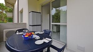 A dining table with plates, cutlery, and a fruit basket, surrounded by chairs, on a balcony with a view of trees and a building. - Pleasant Accommodation With Pool (Lignano Sabbiadoro)