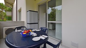 Ein Esstisch mit Tellern, Besteck und einer Obstschale, umgeben von Stühlen, auf einem Balkon mit Blick auf Bäume und ein Gebäude.