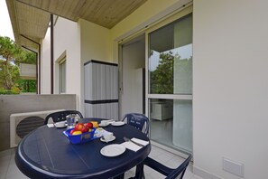 A dining table with plates, cutlery, and a fruit basket, surrounded by chairs, on a balcony with a view of trees and a building. - Relaxing Retreat for a Comfortable Stay (Lignano Sabbiadoro)
