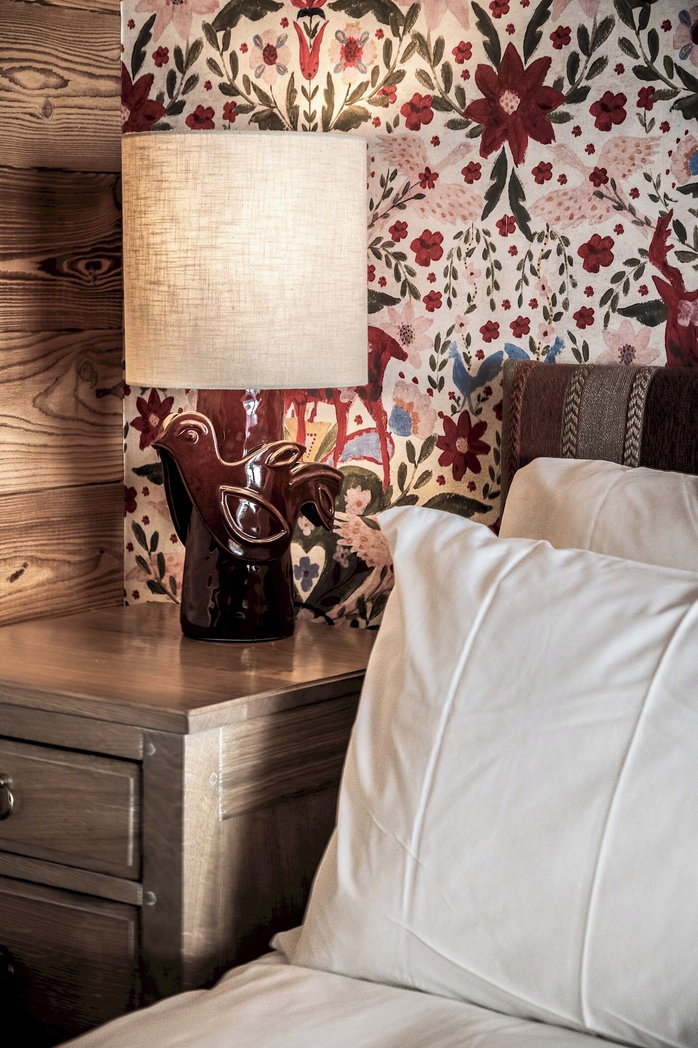 A wooden nightstand with a lamp and a bird-shaped candle holder, a white pillow, and a floral-patterned wall.