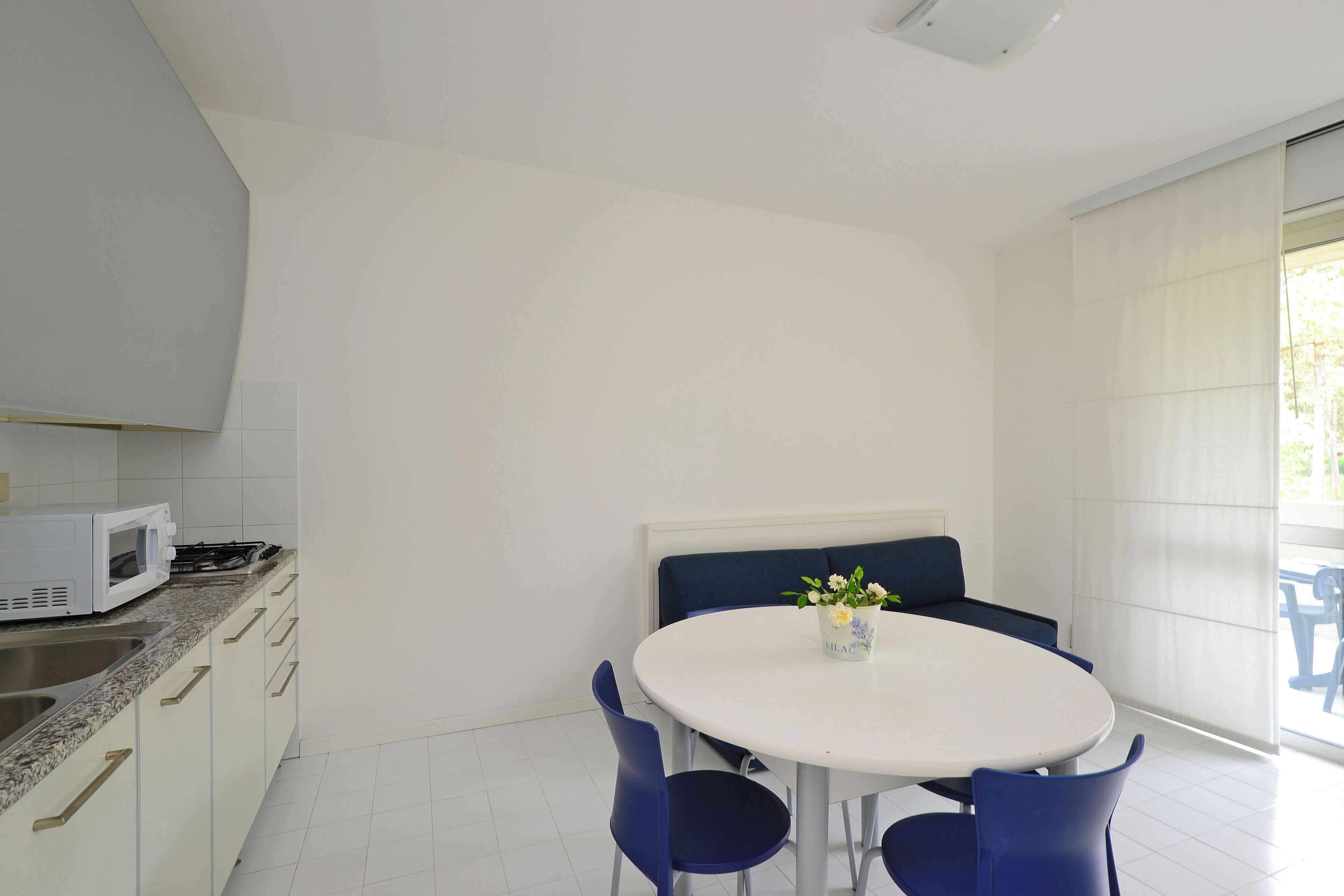 A modern kitchen with white cabinets, a microwave, and a gas stove. There is a round table with blue chairs and a bench with a vase of flowers.