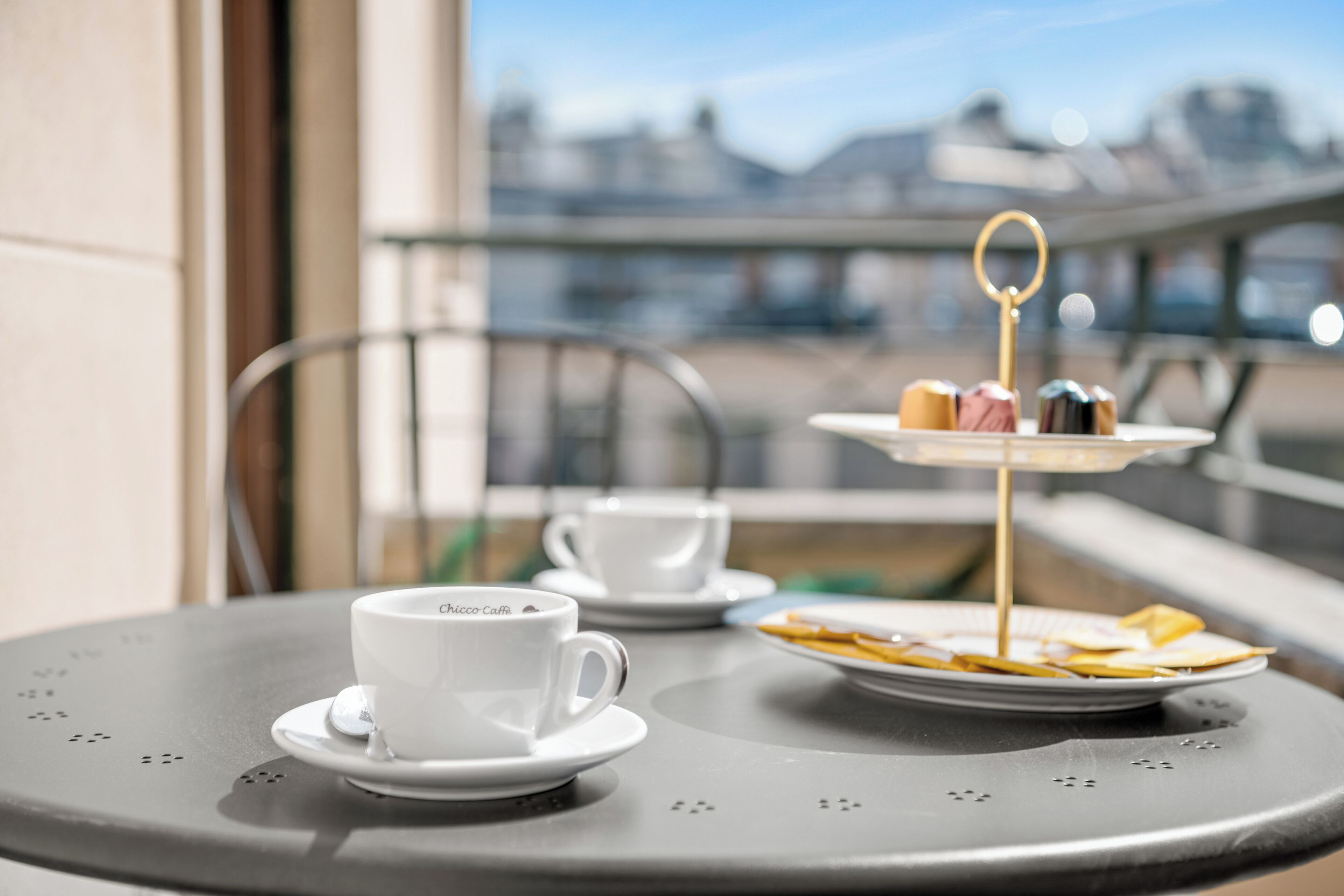 Ein Tisch mit einer weißen Tasse und Untertasse, einem Tablett mit Gebäck und einer Etagere für Desserts auf einem Balkon mit Blick auf die Stadt.