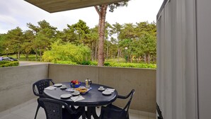 Un espace de restauration en plein air avec une table dressée pour quatre personnes, entourée de chaises, et une vue sur un parc avec des arbres et une route en arrière-plan.