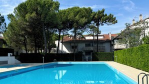 Une piscine dont les bords sont recouverts de carreaux, entourée d’arbres et bordée d’un bâtiment dont le toit est fait de tuiles rouges.