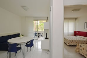 A room with a white desk, blue chairs, a white sofa, and two beds with patterned bedding. - Stylish Flat for a Perfect Getaway in Lignano (Lignano Sabbiadoro)