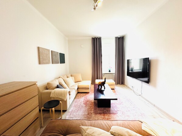 Un salon comprenant un canapé, une table basse, un téléviseur fixé au mur et une fenêtre avec des rideaux.