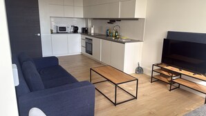 En moderne stue med sofa, sofabord, fjernsyn og et køkken med hårde hvidevarer.
