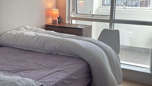 En seng med hvid dyne, et natbord med lampe og et vindue med udsigt til en bygning.