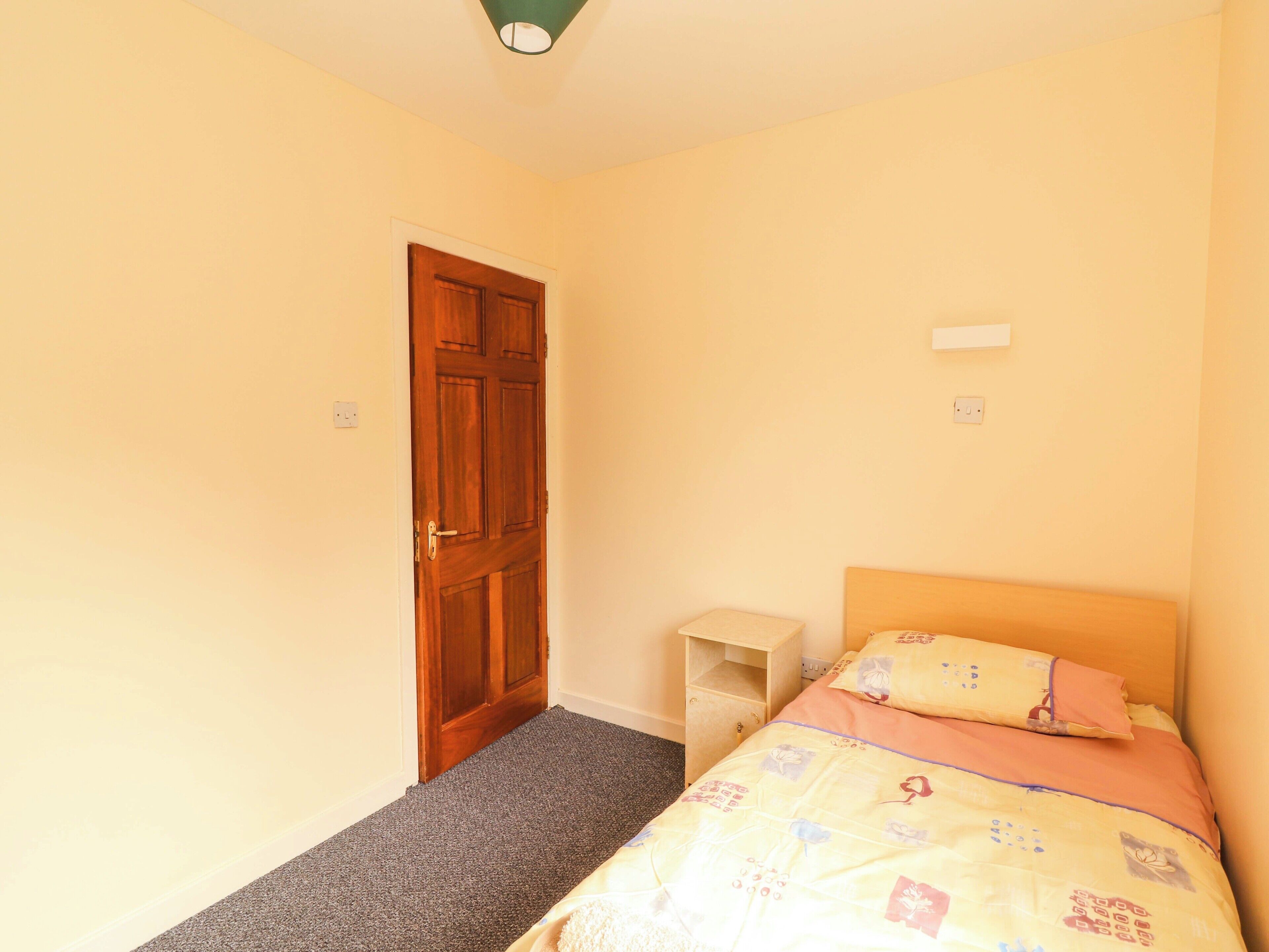 A bedroom with a wooden door, a bed with a patterned bedspread, a small wooden nightstand, and a carpeted floor.