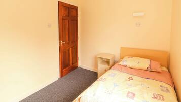 A bedroom with a wooden door, a bed with a patterned bedspread, a small wooden nightstand, and a carpeted floor.