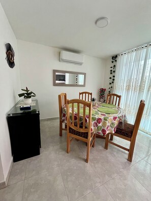 Un comedor con una mesa redonda, sillas de madera, una mesita auxiliar negra, aire acondicionado instalado en la pared, un espejo y una ventana con cortinas.