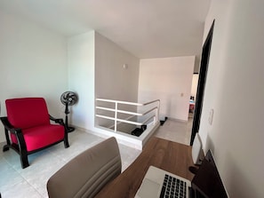 Una sala de estar con una silla roja, una computadora portátil sobre una mesa de madera, una barandilla blanca y un ventilador.