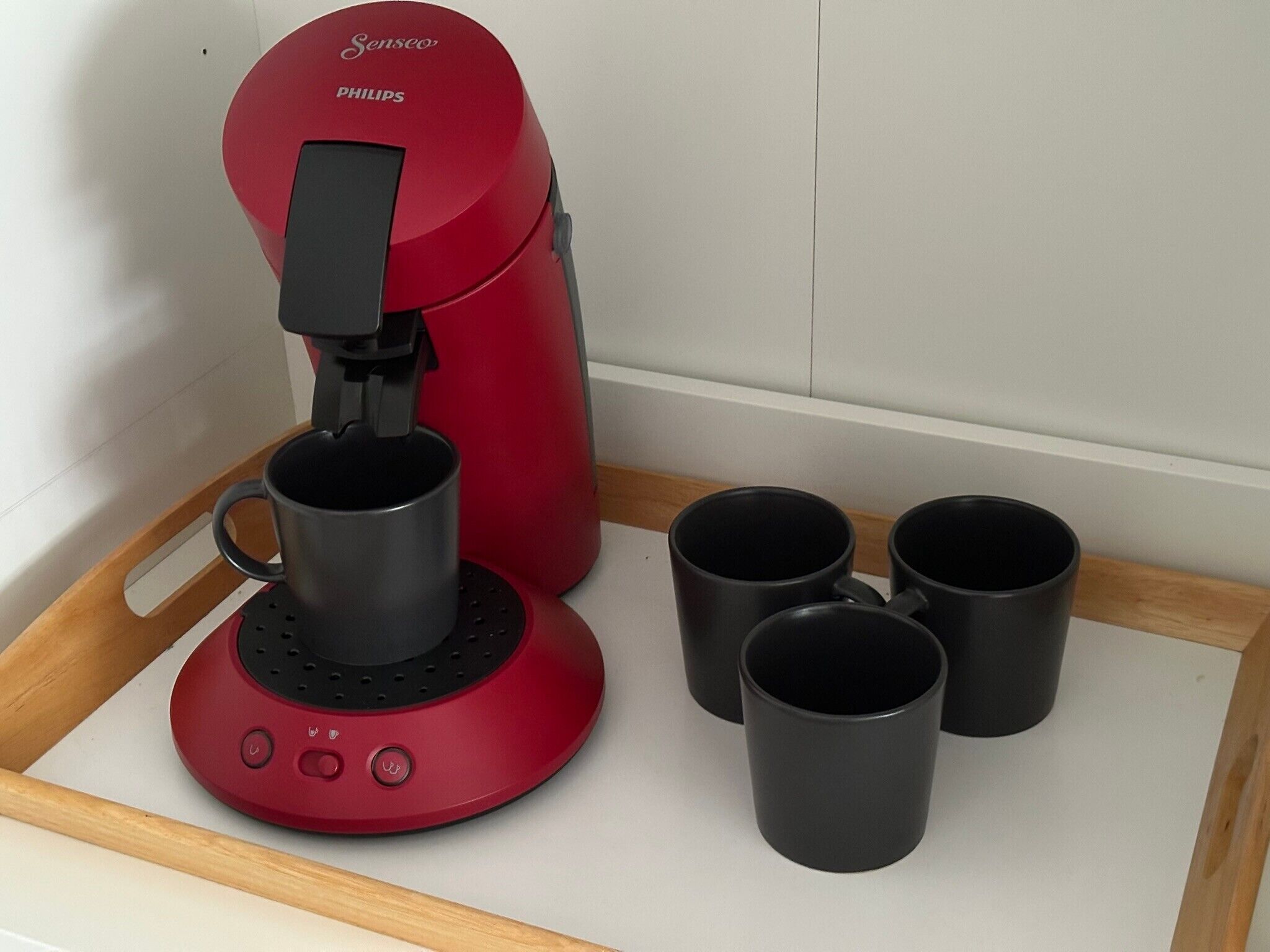 Une machine à café Senseo rouge avec quatre tasses noires posées sur un plateau.