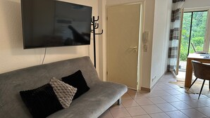 Una sala de estar con un sofá gris, un televisor de pantalla plana montado en la pared, una puerta y vistas al exterior a través de una ventana.