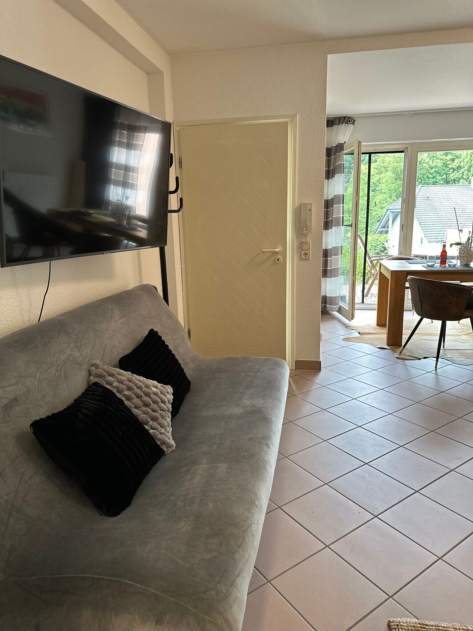 A living room with a sofa, a flat-screen TV mounted on the wall, a door leading to another room, and a view of the outside through a glass door.