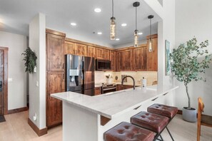 A modern kitchen with wooden cabinets, stainless steel appliances, a large island with bar stools, pendant lighting, and a potted plant. - Remodeled Condo, Ski in/out with Hot Tub, Spa, Gym (Breckenridge)