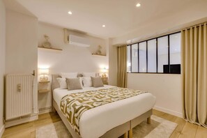 A bedroom with a bed, bedside lamps, a radiator, a window with curtains, and a wall-mounted air conditioner. - A Cosy 2-Br/2BA Duplex - Louvre/Rivoli (Paris)