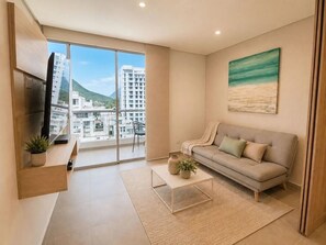 A modern living room with a sofa, coffee table, TV, and a view of buildings and a mountain. - Salguero Sunset Apartment – Near the Sea SOHO-382A (Santa Marta)