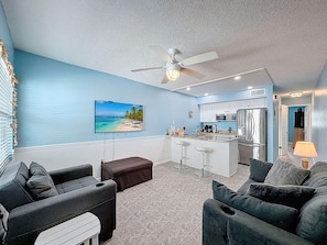 A living room with a TV, a ceiling fan, a kitchen area, and a bar. - Harbor House 26 (Treasure Island)