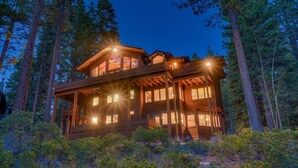 Une cabane en bois à plusieurs étages, dotée de grandes fenêtres et d’un balcon, entourée de grands arbres.