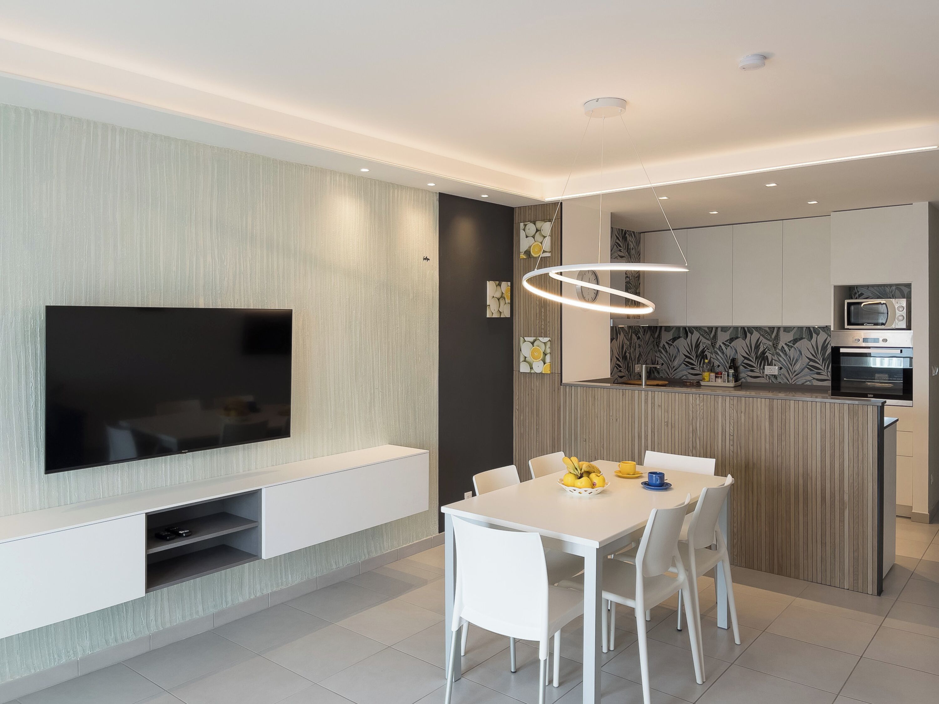 Cozinha moderna com eletrodomésticos embutidos, área de jantar com mesa e cadeiras brancas, e televisão de tela plana fixada na parede.