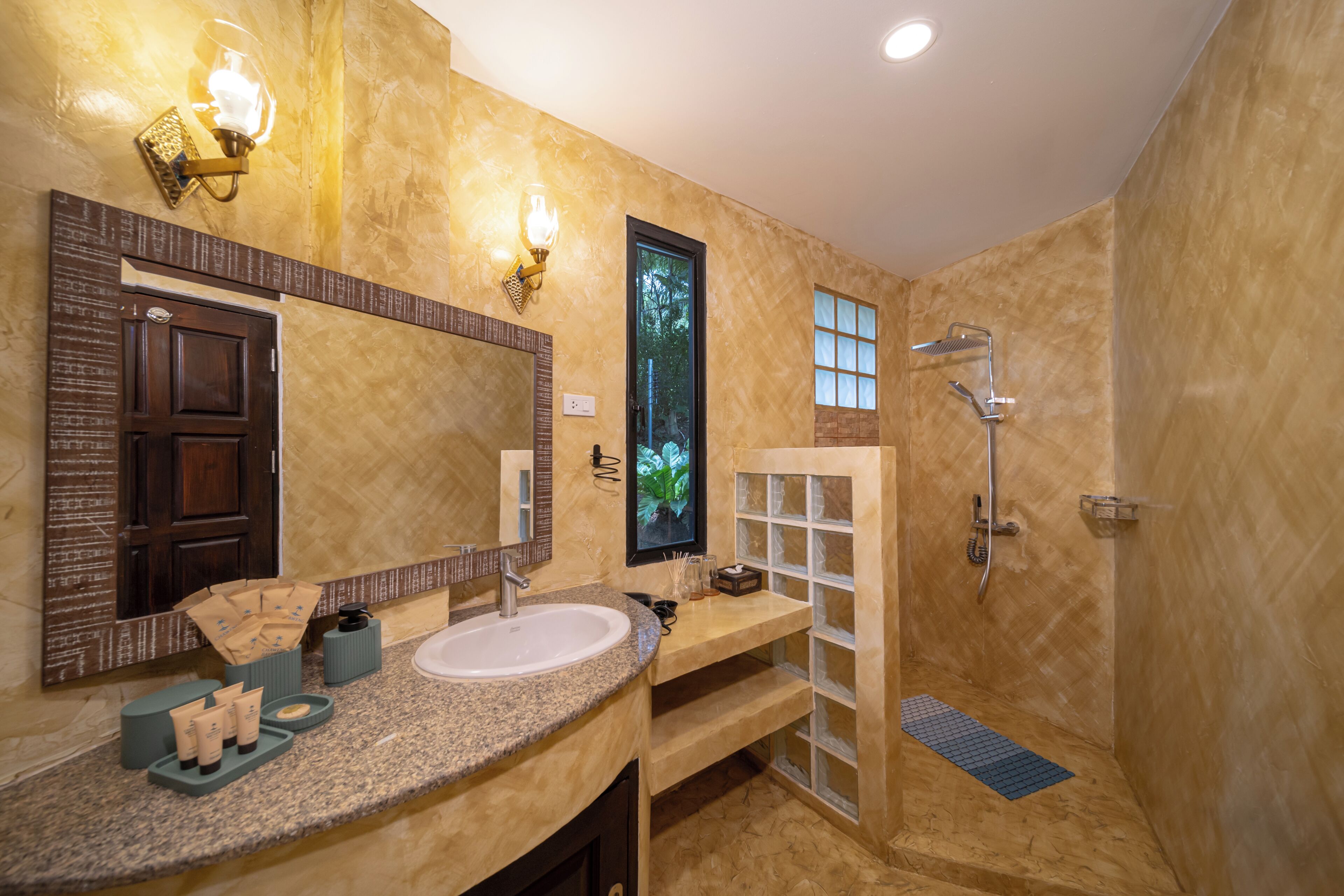 A bathroom with a granite countertop, a large mirror, a shower, and a window with a view of greenery.