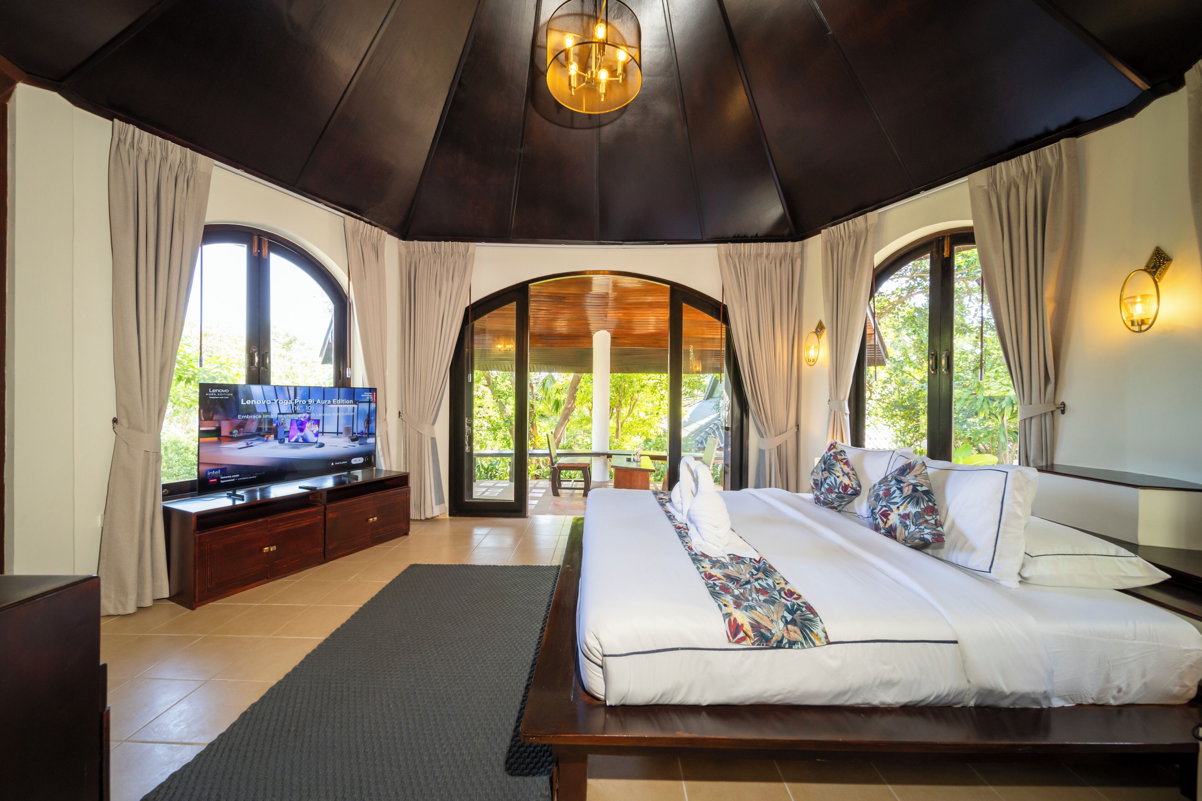 A bedroom with a large bed, a television, and a view of greenery through the windows.