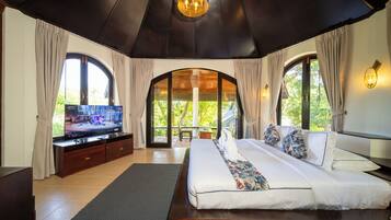 A bedroom with a large bed, a television, and a view of greenery through the windows.