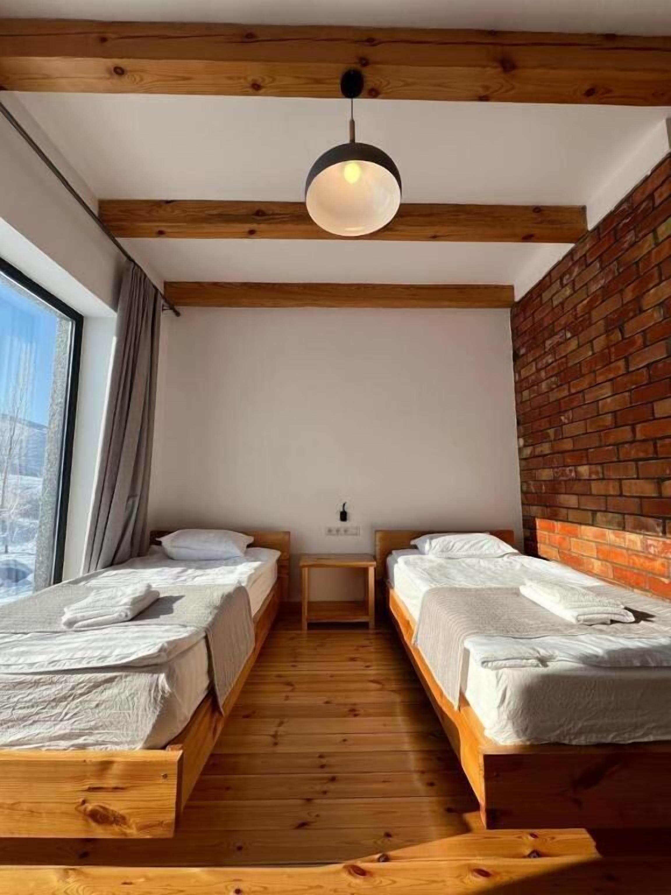 A room with two wooden beds, a small wooden nightstand, a hanging light fixture, and a brick wall.