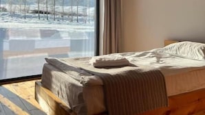 A bedroom with a large window, wooden beams, a bed, and a view of a snowy landscape. - Kemin Guest House (Kaindy)