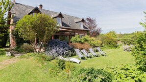 Ein Haus mit geneigtem Dach, Schornstein und einem Garten mit Liegestühlen und Tisch.