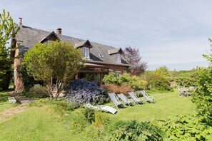Une maison avec un toit en pente, une cheminée et un jardin agrémenté de chaises longues et d’une table.