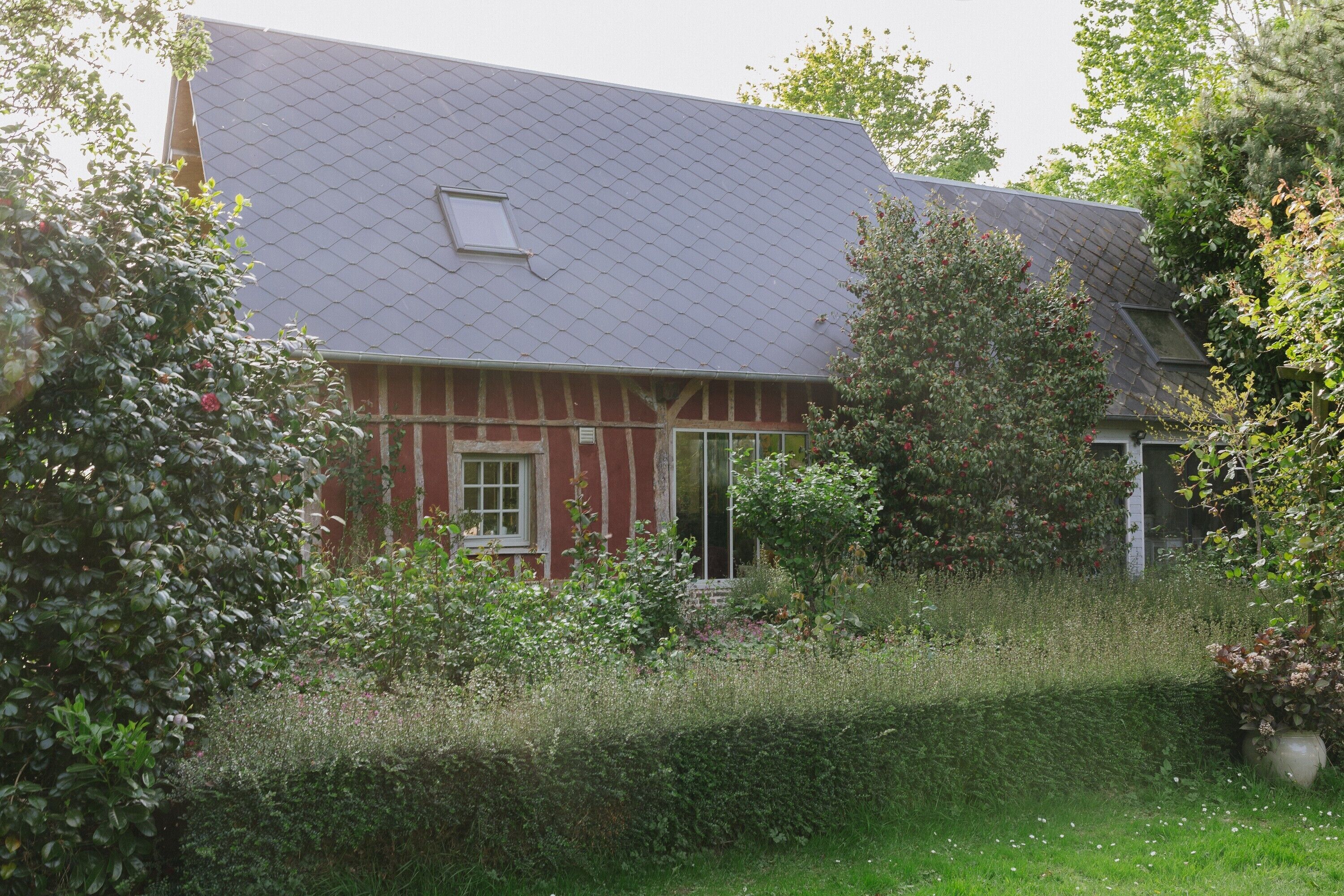 Una casa con fachada roja, techo gris y un jardín con diversas plantas y un jarrón grande.