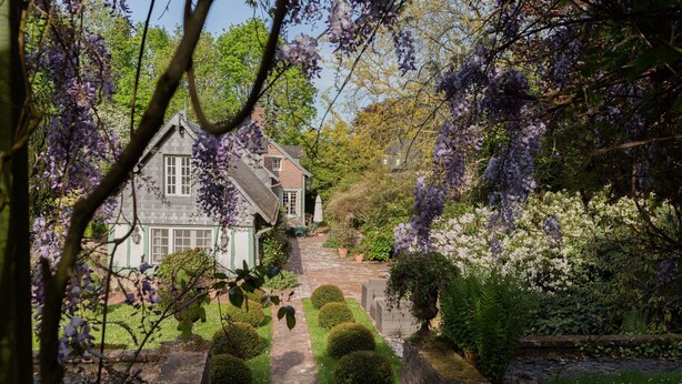 Une maison traditionnelle, avec un chemin de pierre menant à l’entrée, entourée d’une végétation luxuriante et d’arbres en fleurs.