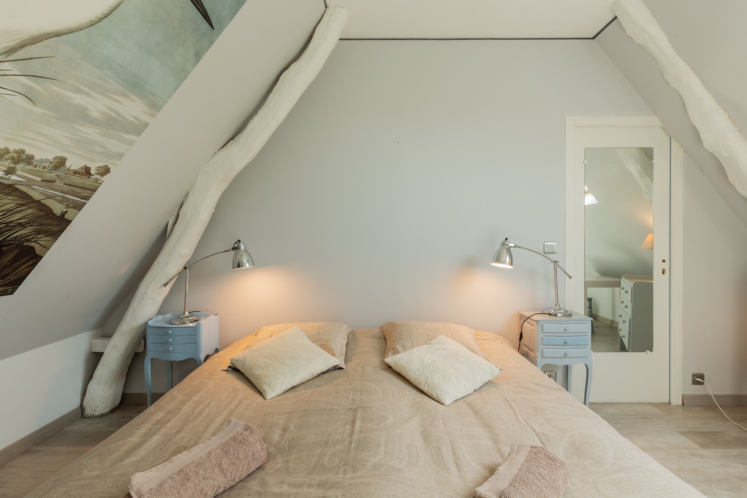 Une chambre avec un plafond en pente, un lit avec deux oreillers, deux tables de chevet avec des lampes et une commode avec un miroir.