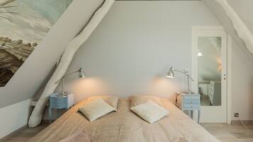 Une chambre avec un plafond en pente, un lit avec deux oreillers, deux tables de chevet avec des lampes et une commode avec un miroir.