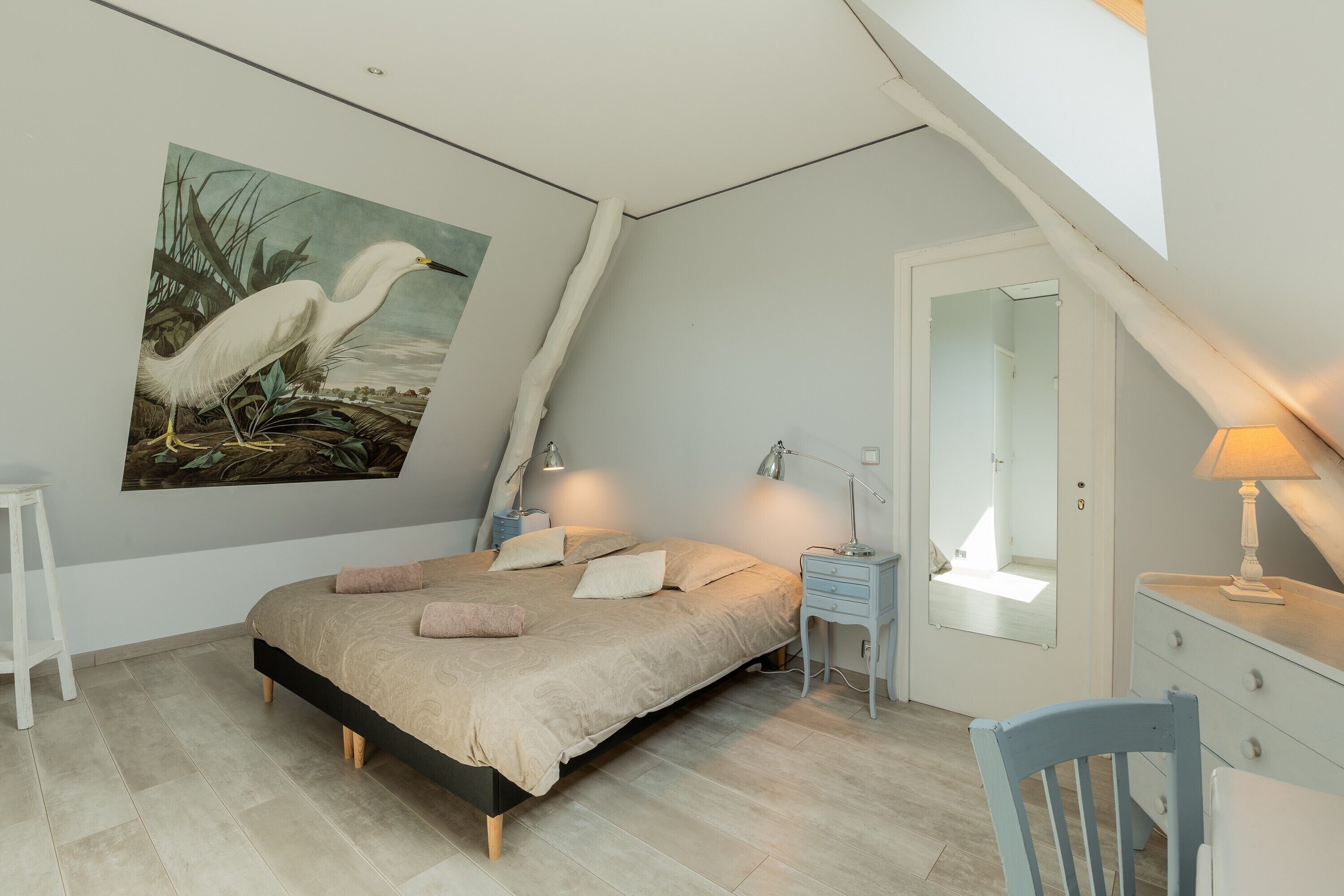 A bedroom with a bed, bedside tables, a chair, a mirror, and a painting of birds.