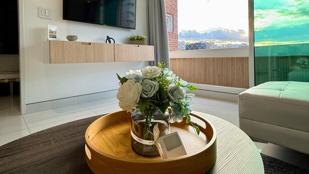 Ein modernes Wohnzimmer mit einem Flachbildfernseher, einem Couchtisch mit Blumenvase, einer Couch und einem großen Fenster mit Blick auf die Stadt.