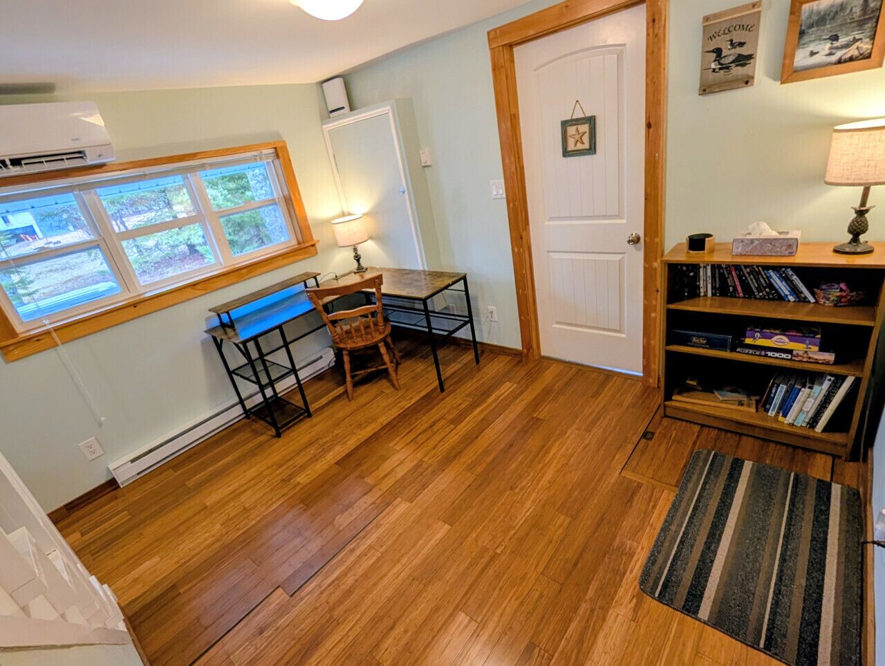 A room with a wooden floor, a desk with a chair, a bookshelf, a door, and a window with blinds.