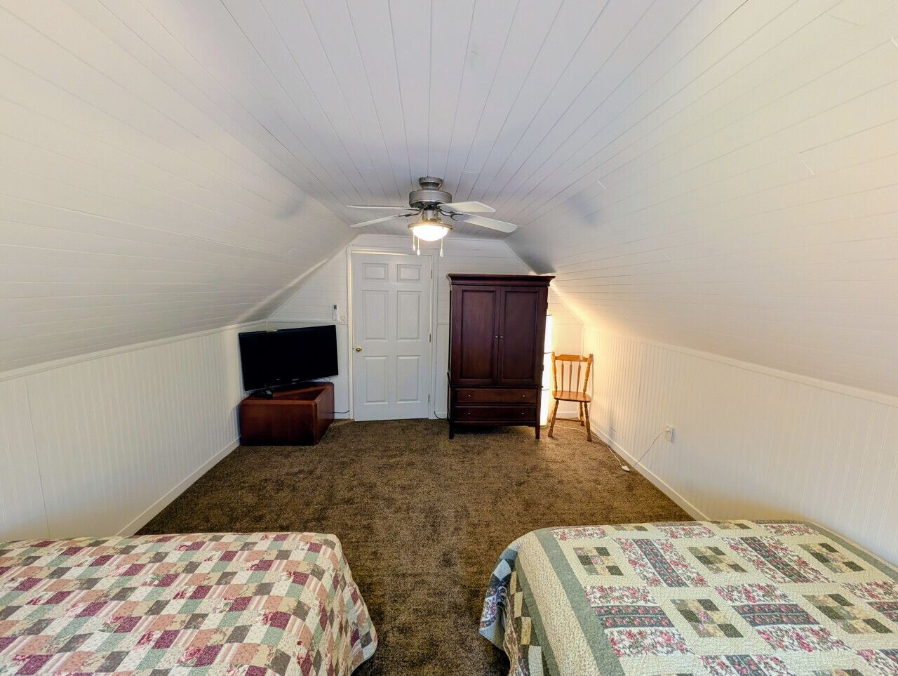 A small, single-room attic bedroom with a sloped ceiling, a ceiling fan, a television on a wooden stand, a wardrobe, and two beds with quilts.