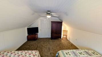 A small, single-room attic bedroom with a sloped ceiling, a ceiling fan, a television on a wooden stand, a wardrobe, and two beds with quilts.