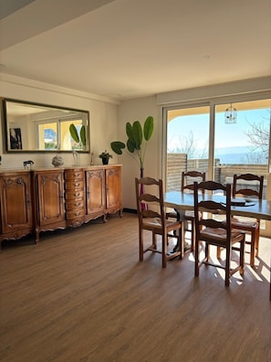 Un coin repas avec une table et des chaises en bois, un grand miroir et une vue à travers une porte-fenêtre coulissante.