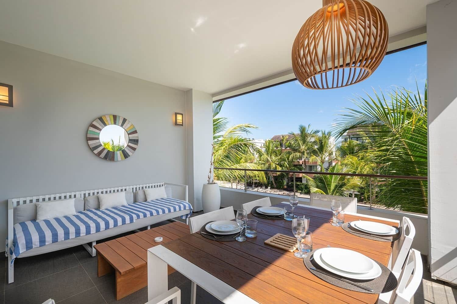 A balcony with a wooden table set for dining, a white sofa with striped cushions, and a large window offering a view of palm trees and buildings.