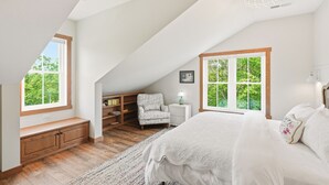 A bedroom with a large bed, a bench, a chair, a nightstand, and a window with a view of greenery.