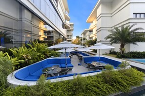 Een modern terras buiten met blauwe zitplaatsen, tafels en parasols, omringd door weelderig groen en een zwembad.
